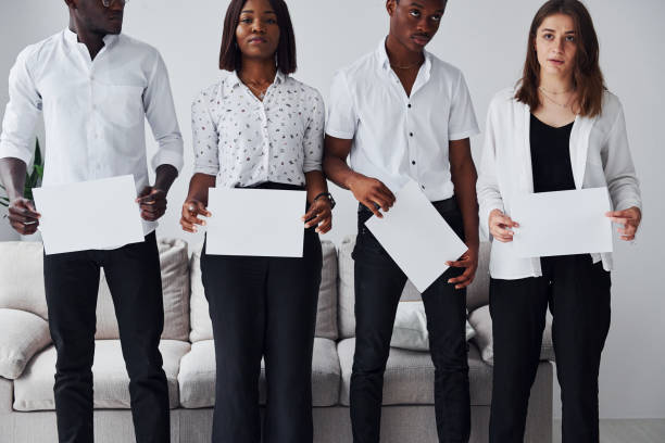 Group of african american people in formal clothes standing near sofa indoors in office together and holding empty papers in hands.